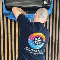 Climatix - Serwisant w koszulce firmowej ClimatiX dokonuje przeglądu klimatyzacji, zabezpieczonej niebieskim pokrowcem, na tle ściany z drewnianymi panelami.