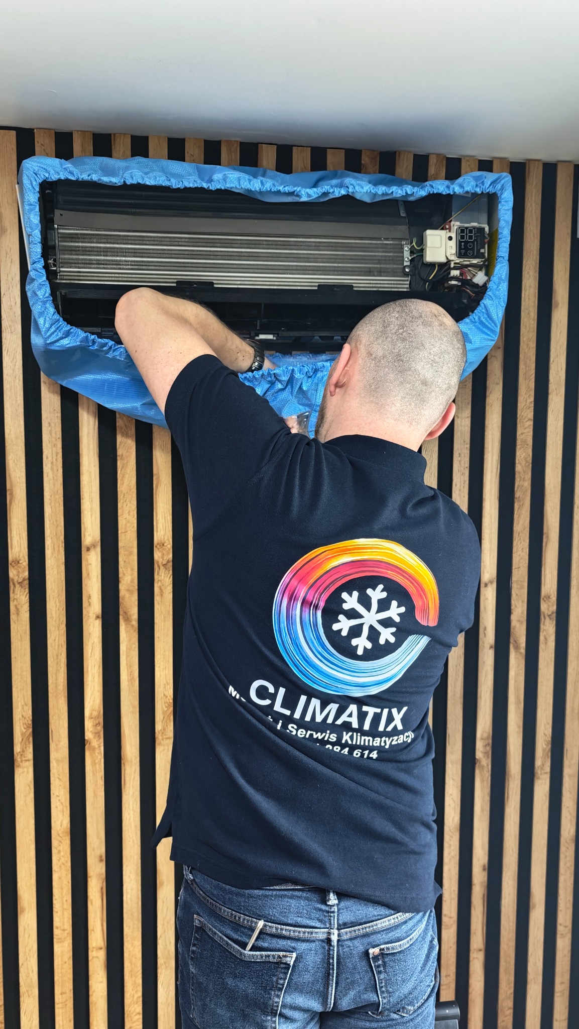Serwisant w koszulce firmowej ClimatiX dokonuje przeglądu klimatyzacji, zabezpieczonej niebieskim pokrowcem, na tle ściany z drewnianymi panelami.
