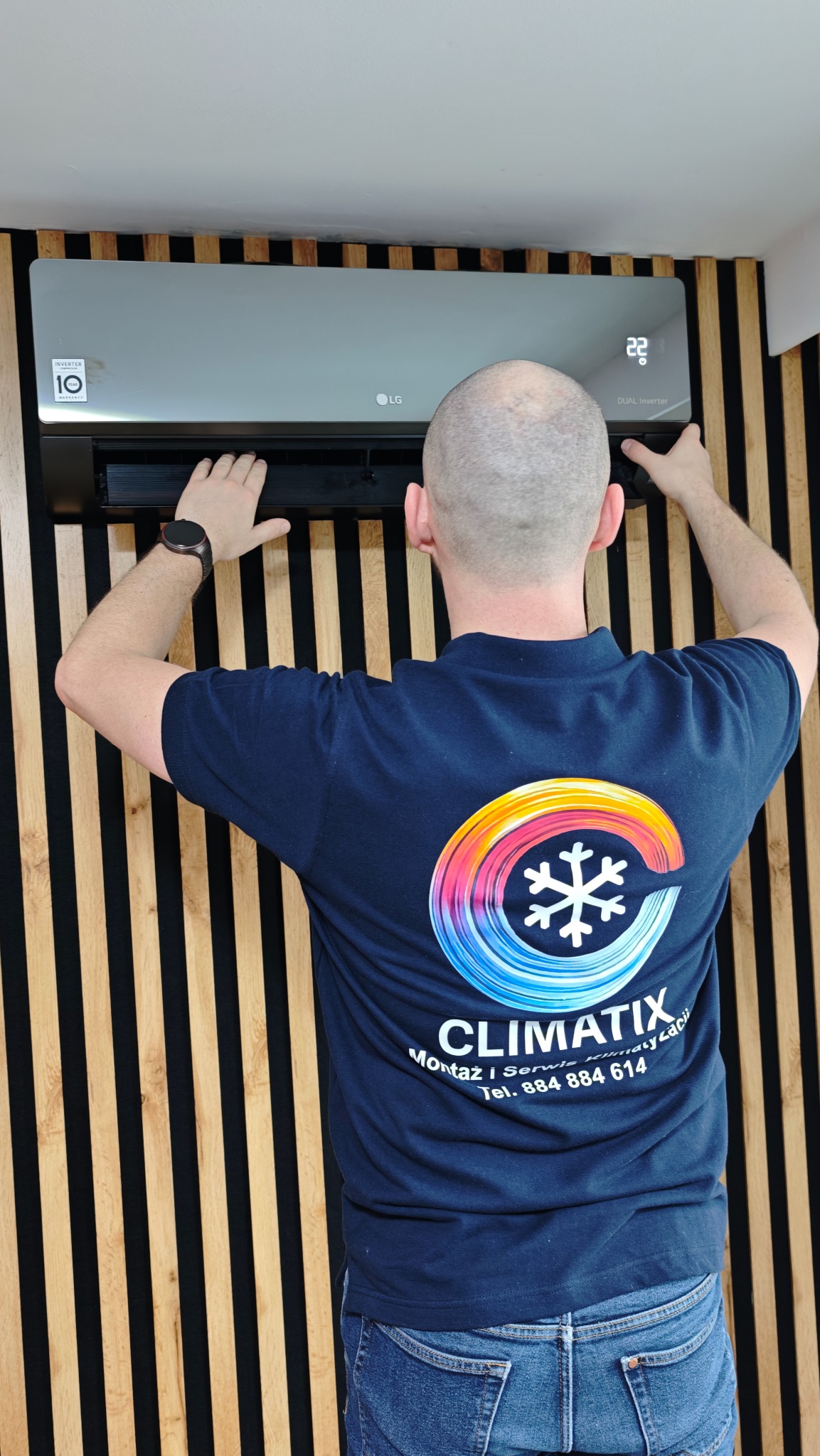 Przegląd klimatyzacji LG Dual Inverter na ścianie z drewnianymi panelami. Mężczyzna w koszulce firmowej ClimatiX wykonuje serwis. Widoczny wyświetlacz z temperaturą 22°C.