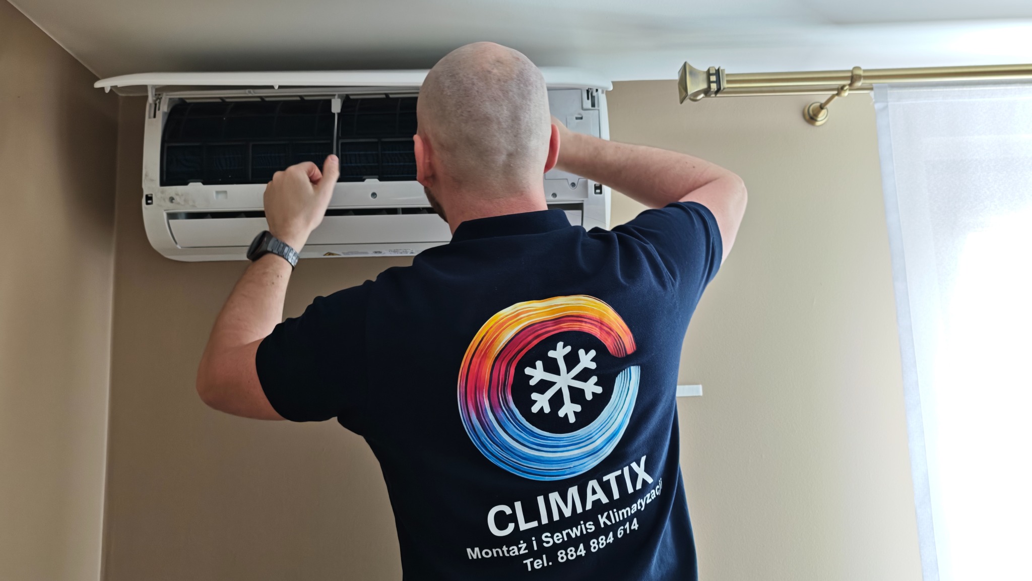 Serwisant w firmowej koszulce z logo ClimatiX, podczas przeglądu klimatyzacji ściennej w jasnym wnętrzu. Widoczne filtry urządzenia i fragment zasłony.