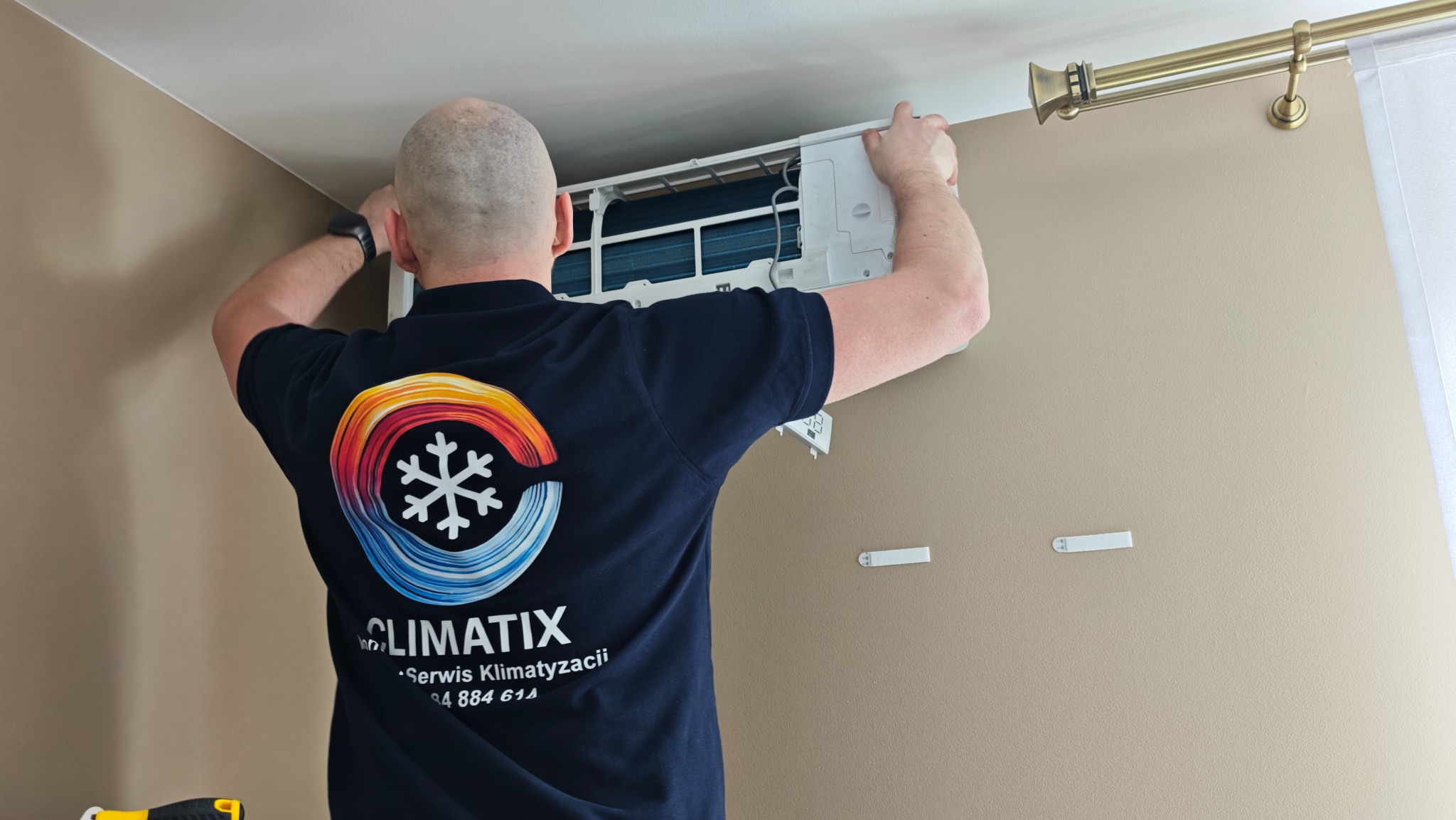 Serwisant w koszulce firmowej montuje klimatyzator na ścianie w pomieszczeniu. Widoczne elementy instalacji i narzędzia.
