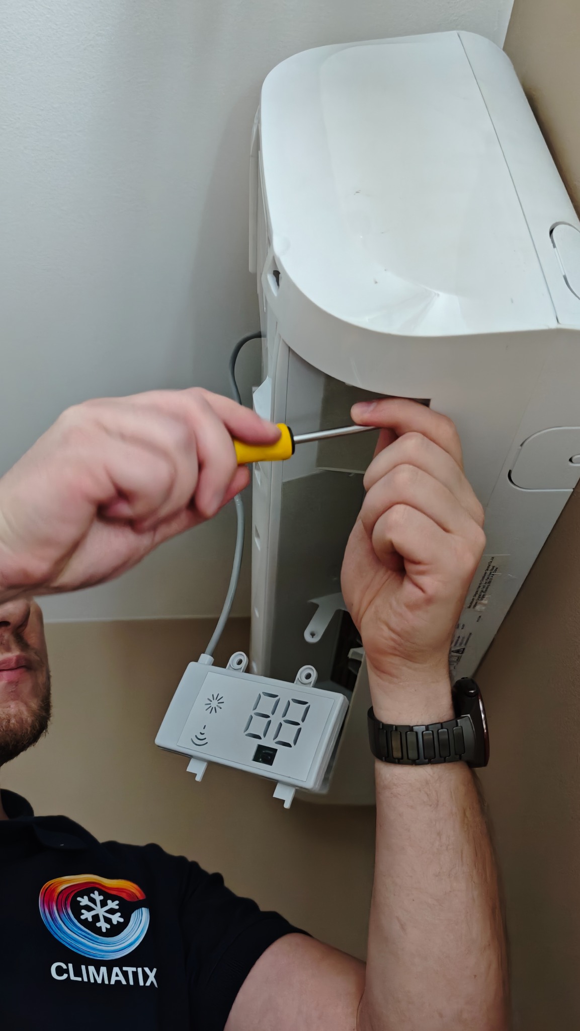 Serwisant z żółtym śrubokrętem dokonuje przeglądu klimatyzacji ściennej. Widoczny panel sterowania z wyświetlaczem i logo firmy na koszulce serwisanta.
