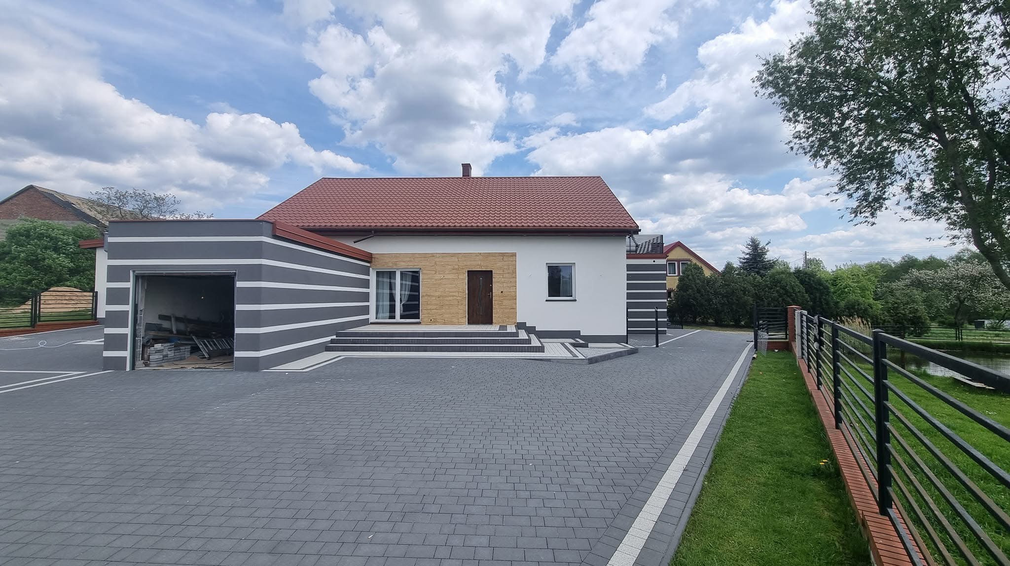 Nowoczesny dom z garażem w szaro-białe pasy. Elewacja z elementami dekoracyjnymi, kostka brukowa na podjeździe, ogrodzenie. Słoneczny dzień.