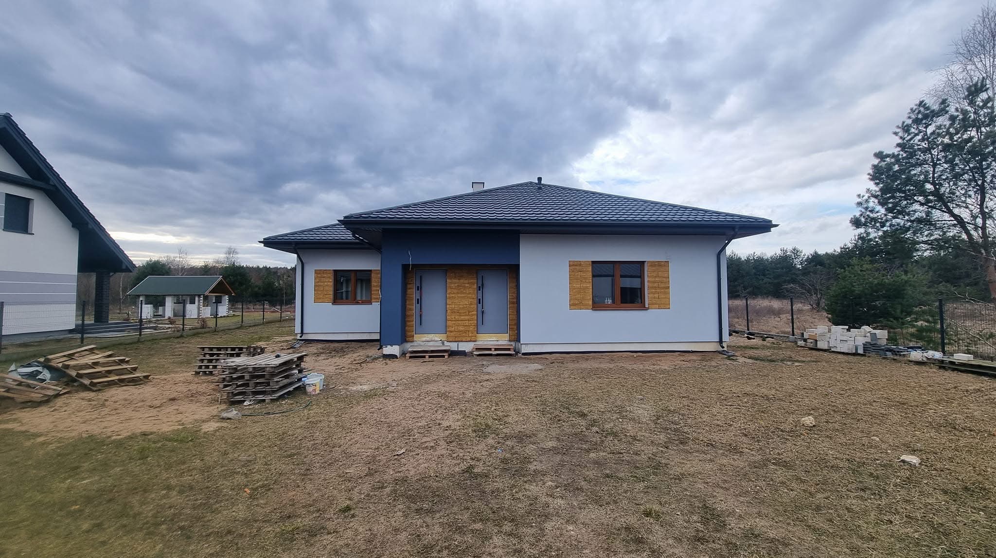 Nowoczesny parterowy dom z szarą elewacją, drewnianymi detalami okien i niebieskim akcentem przy wejściu, otoczony posesją w trakcie prac wykończeniowych.