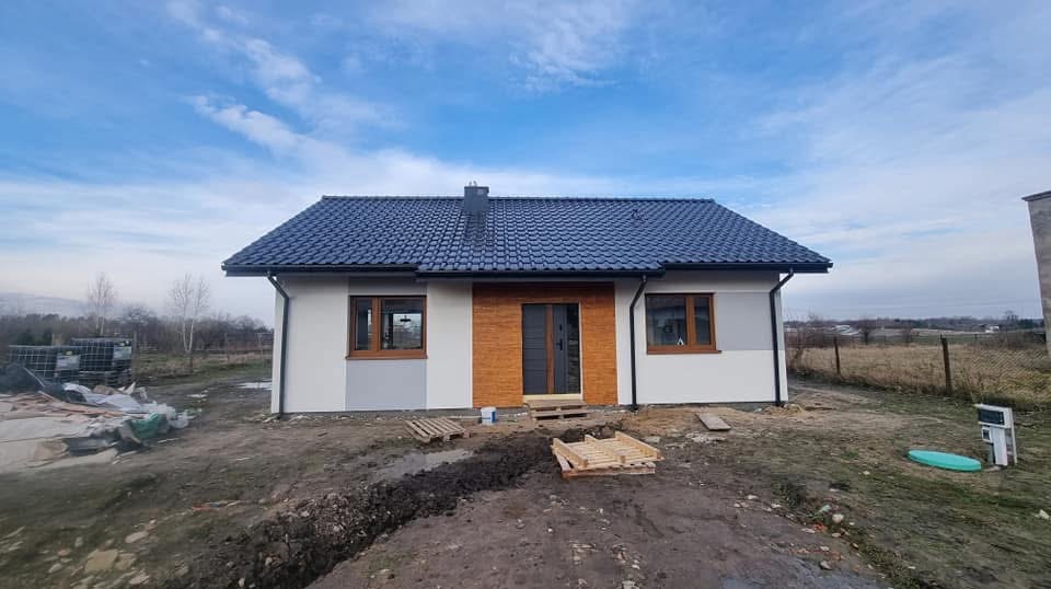 Nowoczesny, parterowy dom z białą elewacją, drewnianymi akcentami i ciemną dachówką na tle błękitnego nieba. Uwagę przykuwają palety przed wejściem.