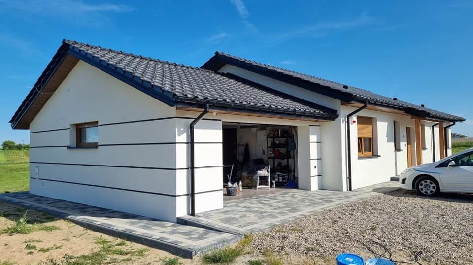 Biały dom parterowy z garażem, ciemna dachówka, elewacja z poziomymi czarnymi pasami. Kostka brukowa przed garażem, fragment samochodu z boku.