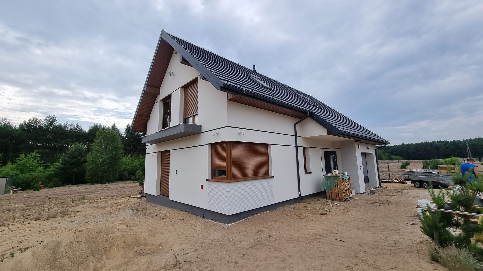 Nowoczesny dom jednorodzinny z białymi ścianami, brązowymi roletami i ciemną dachówką, widoczny z boku na tle piaszczystego terenu i zieleni.