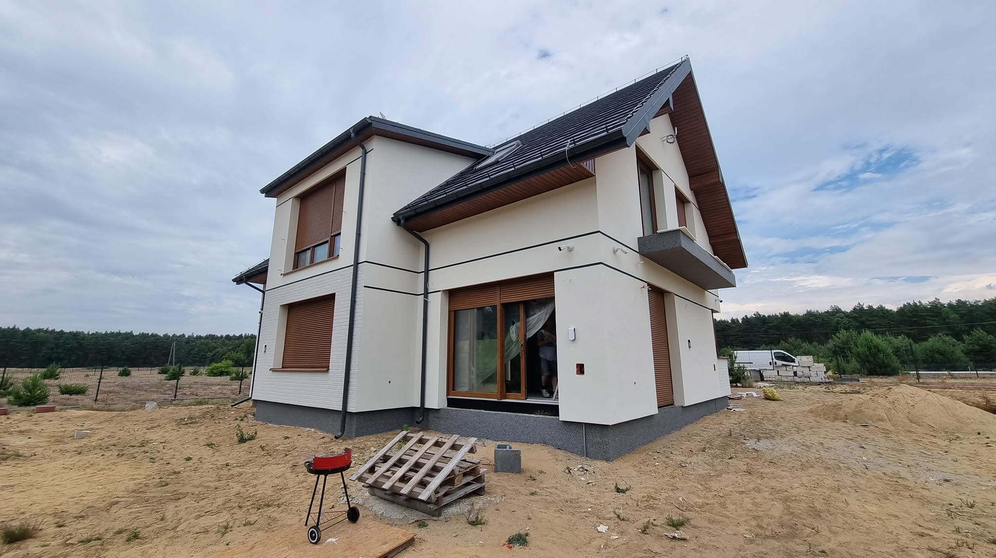 Dwukondygnacyjny dom z jasną elewacją, brązowymi roletami i ciemnym dachem. Widoczny grill i paleta. Tło stanowią drzewa i pochmurne niebo.