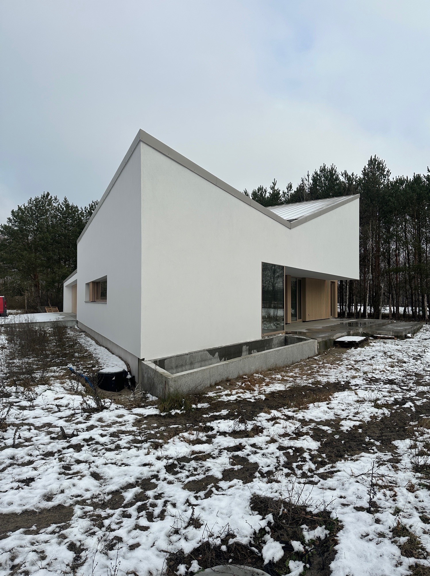 Nowoczesny dom z białą elewacją i szarym dachem na tle lasu w zimowej scenerii. Widoczne okna i betonowe elementy w otoczeniu. Minimalistyczny design.