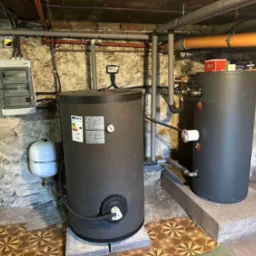 Dwa zbiorniki akumulacyjne w piwnicy z kamiennymi ścianami w Józefosławiu, obok skrzynka elektryczna i instalacja hydrauliczna, widoczne rury i podłączenia.