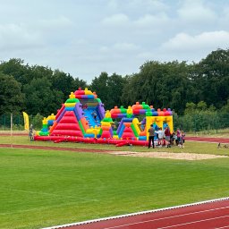 RAKPOL PAWEŁ RACZYŃSKI - Kolorowy, dmuchany plac zabaw na zielonym boisku sportowym. W tle drzewa i pochmurne niebo. Dzieci bawią się na dmuchańcu.