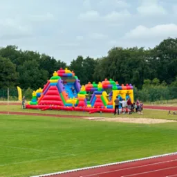 Kolorowy, dmuchany plac zabaw na zielonym boisku sportowym. W tle drzewa i pochmurne niebo. Dzieci bawią się na dmuchańcu.
