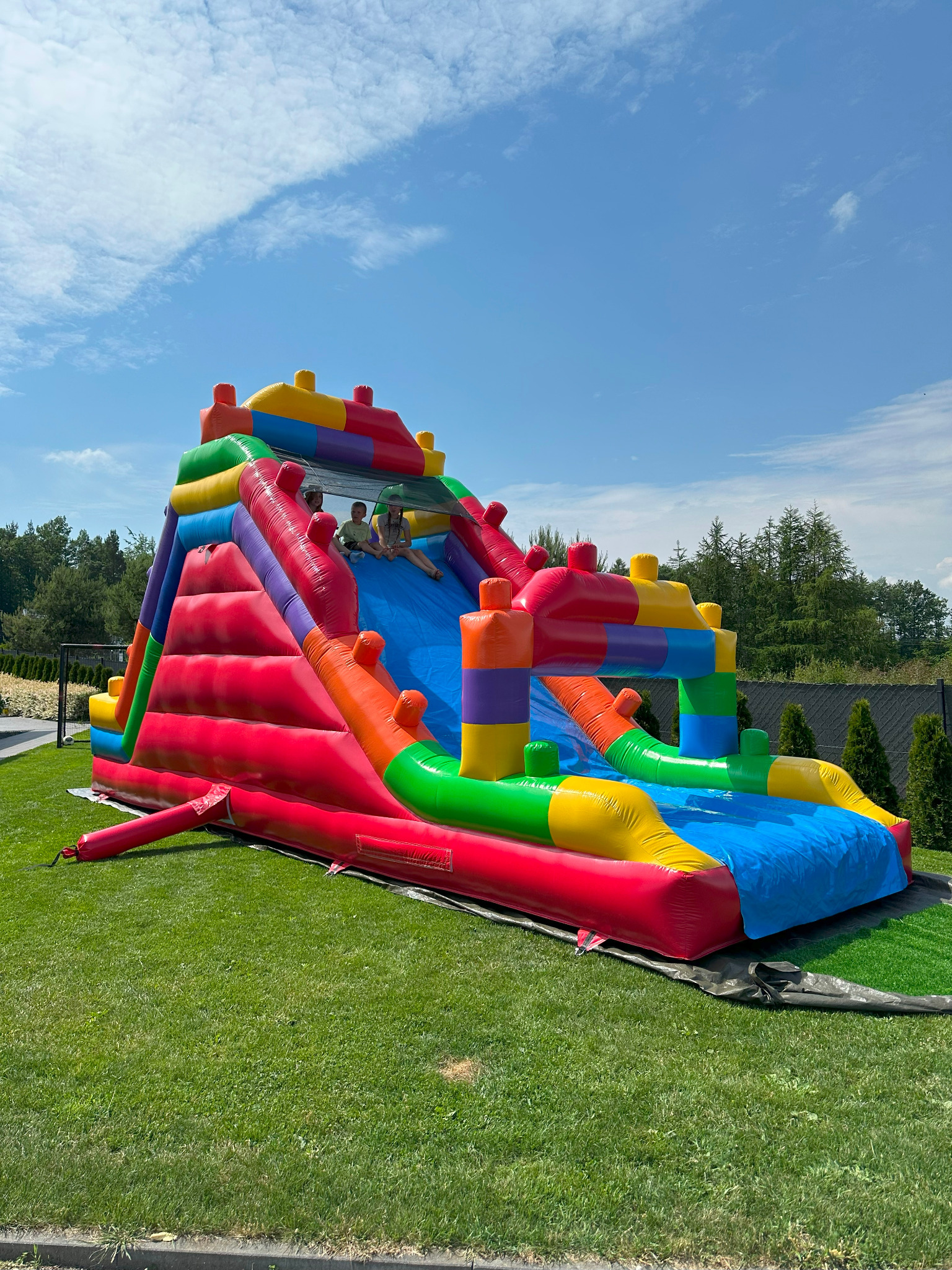 Kolorowa, dmuchana zjeżdżalnia na trawie w słoneczny dzień. Dzieci siedzą na górze zjeżdżalni. W tle widoczne drzewa i niebieskie niebo.