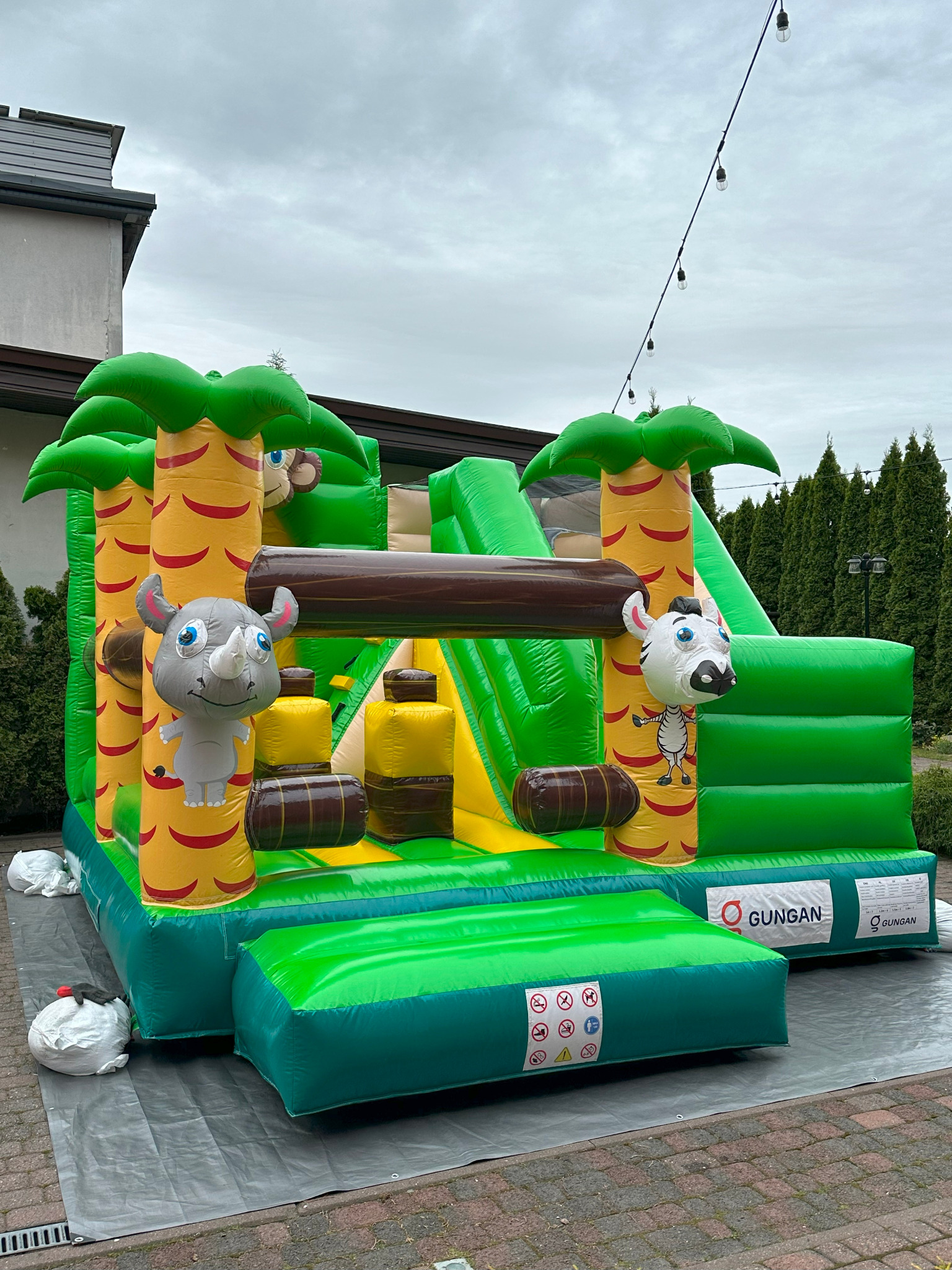 Kolorowy dmuchany zamek z motywem dżungli: palmy, małpa, zebra i nosorożec. Zamek stoi na szarej macie, w tle budynek i tuje. Oświetlenie dekoracyjne nad zamkiem.