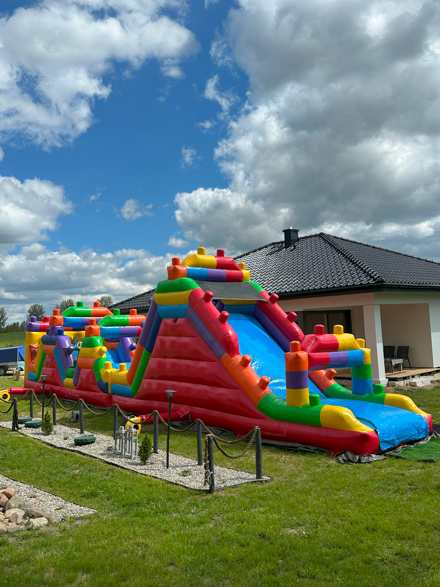 Kolorowy, dmuchany tor przeszkód na trawniku przed domem z czarnym dachem, błękitne niebo z chmurami w tle. Atrakcja na imprezę plenerową.