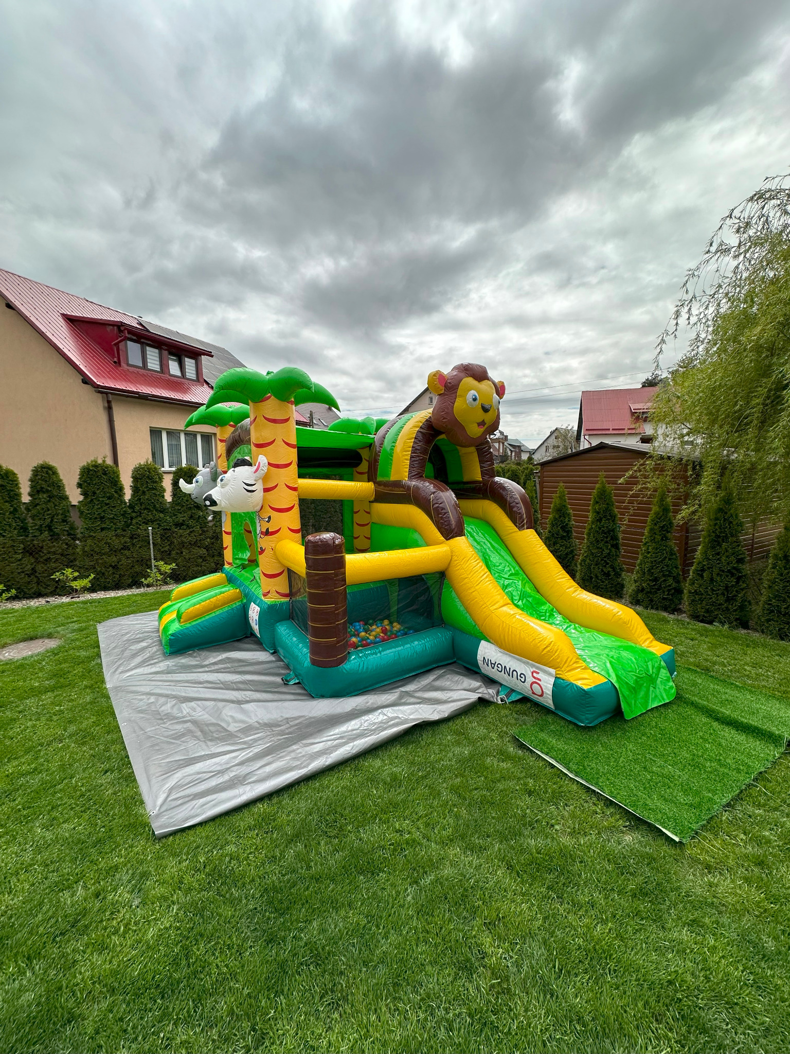 Kolorowy, dmuchany plac zabaw w dżungli z motywami zwierzęcymi, rozłożony na trawniku. Widoczne elementy zjeżdżalni i basenu z kulkami.