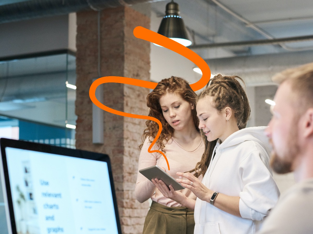 Dwie młode kobiety analizują dane na tablecie w nowoczesnym biurze. Widoczny monitor z wykresami, ceglany filar i lampa sufitowa. Współpraca zespołu w jasnym otoczeniu.