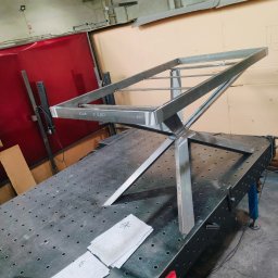 Tomasz Bierowiec - Konstrukcja metalowa z profili na stole spawalniczym z rysunkami technicznymi i narzędziami. Widoczne spawy i oznaczenia na metalu. Surowy stan, warsztatowy klimat.