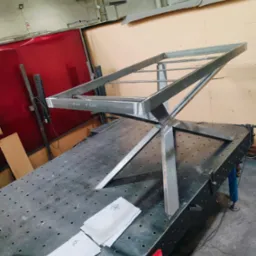 Konstrukcja metalowa z profili na stole spawalniczym z rysunkami technicznymi i narzędziami. Widoczne spawy i oznaczenia na metalu. Surowy stan, warsztatowy klimat.