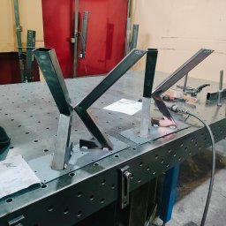 Tomasz Bierowiec - Spawanie metalowych elementów konstrukcyjnych na stole spawalniczym. Widoczne spawy, narzędzia ślusarskie i rysunek techniczny. Elementy w trakcie spawania.