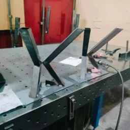 Spawanie metalowych elementów konstrukcyjnych na stole spawalniczym. Widoczne spawy, narzędzia ślusarskie i rysunek techniczny. Elementy w trakcie spawania.