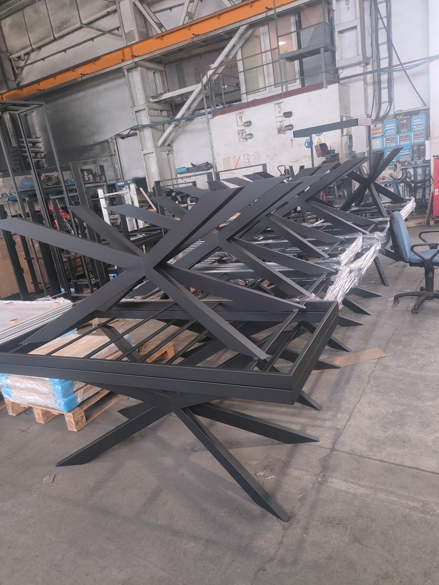 Stalowe, pomalowane proszkowo konstrukcje nóg do stołów ułożone na palecie w hali produkcyjnej. Widoczne spawy i elementy mocujące. Surowe, przemysłowe otoczenie.