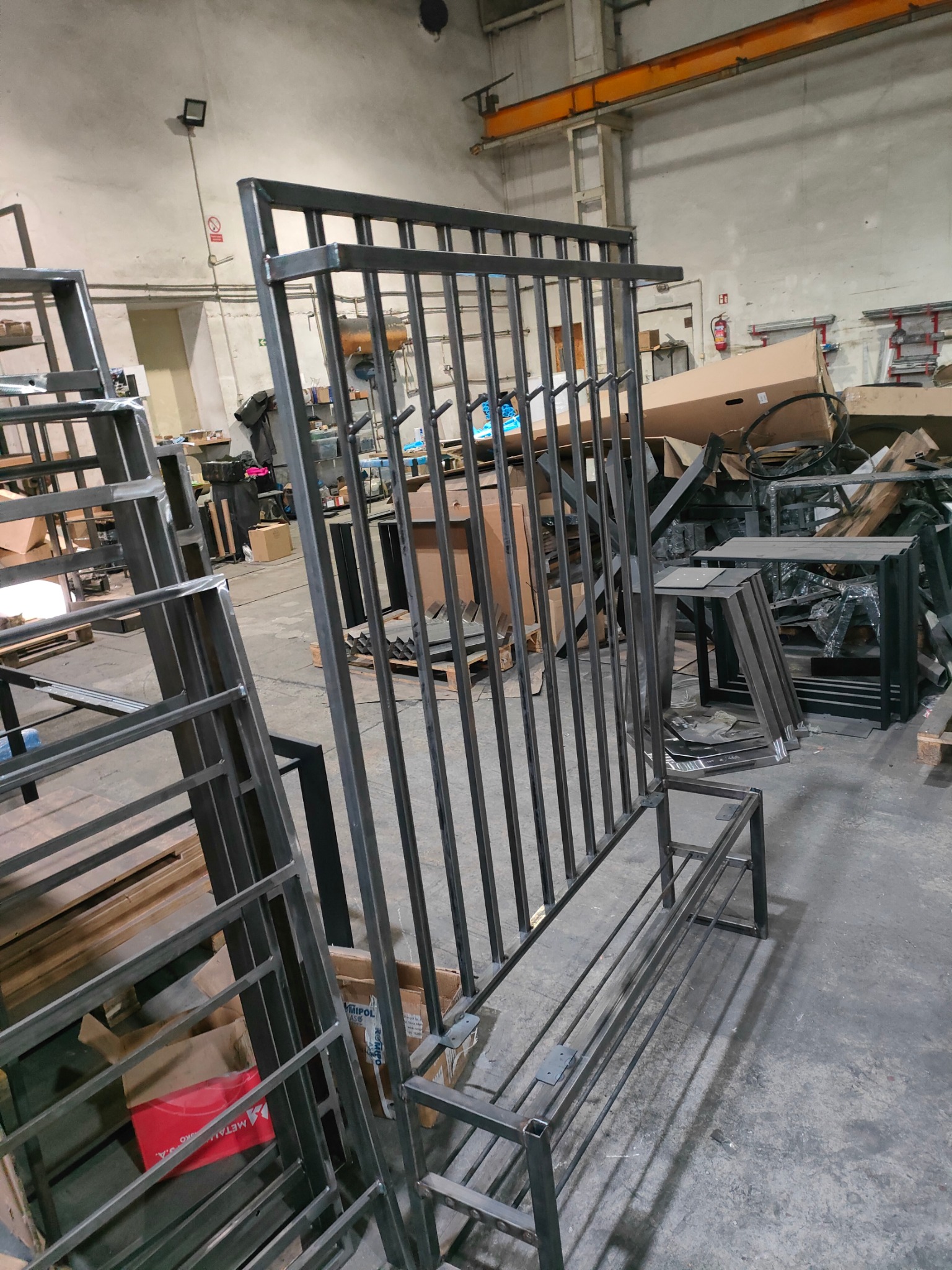 Metalowa konstrukcja z pionowymi prętami w warsztacie, surowy stan, widoczne spawy, elementy w trakcie produkcji, przemysłowe otoczenie.