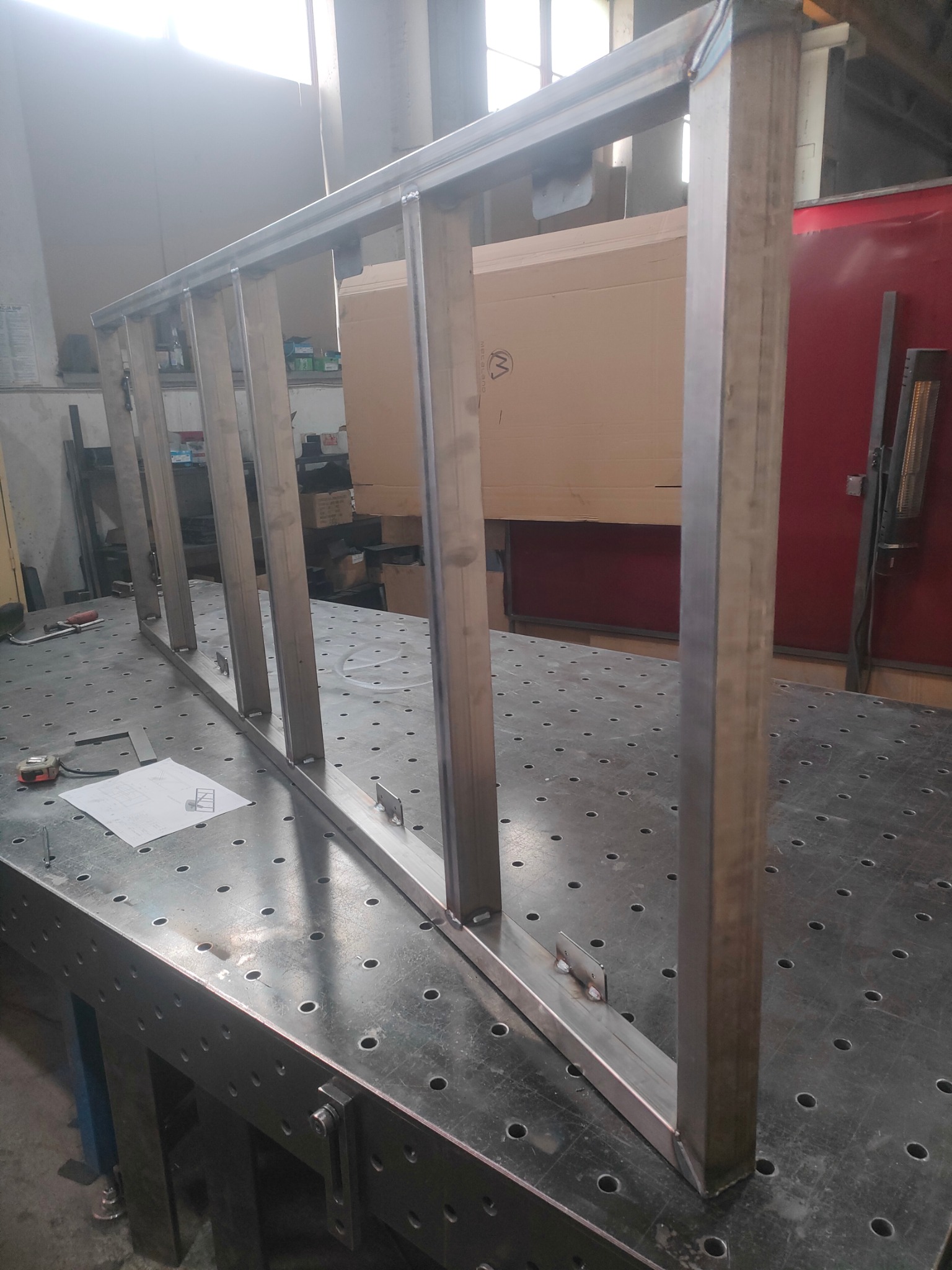 Konstrukcja metalowa z profili, spawana na stole ślusarskim, widoczne spawy, plan techniczny obok. Element konstrukcyjny w trakcie tworzenia w warsztacie.