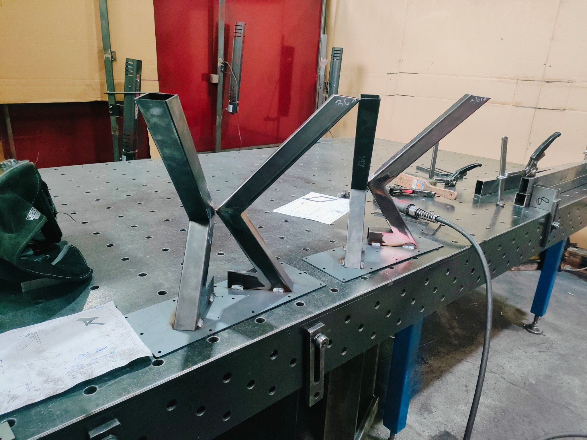 Spawanie metalowych elementów konstrukcyjnych na stole spawalniczym. Widoczne spawy, narzędzia ślusarskie i rysunek techniczny. Elementy w trakcie spawania.