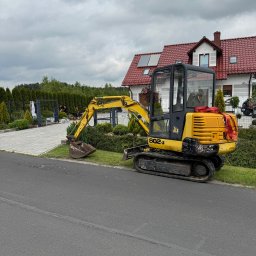 ZIELONA PRZESTRZEŃ MARTYNA SEJUD - Żółta mini koparka JCB 802.4 stoi na poboczu drogi asfaltowej, w tle dom z czerwoną dachówką i pracownicy przy ogrodzeniu z roślinami. Pochmurne niebo.