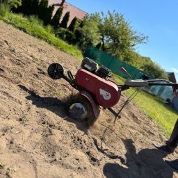 ZIELONA PRZESTRZEŃ MARTYNA SEJUD - Czerwony glebogryzarek spalinowy przygotowuje ziemię pod uprawę na działce w słoneczny dzień. W tle widoczny dom z czerwonym dachem i zielone drzewa.