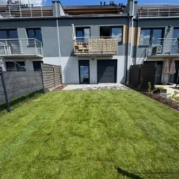 Nowo założony trawnik z rolki przed szeregowcem, z widocznymi elementami systemu nawadniania. Estetyczne wykończenie rabat i ogrodzenia. Słoneczny dzień.