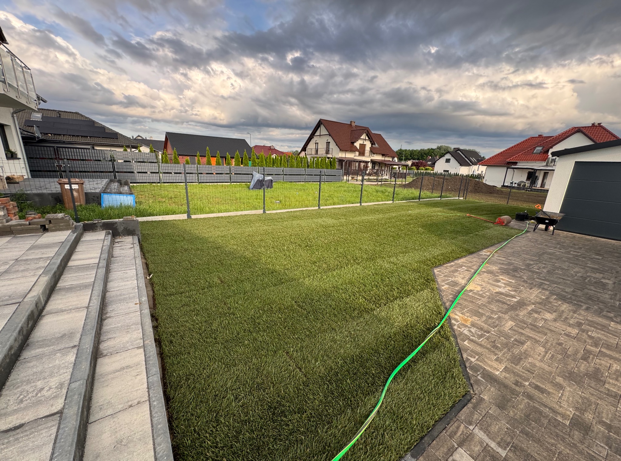 Świeżo ułożona trawa z rolki na posesji, widok z góry. Gęsta, zielona darń kontrastuje z szarą kostką brukową i ogrodzeniem. Zachmurzone niebo w tle.