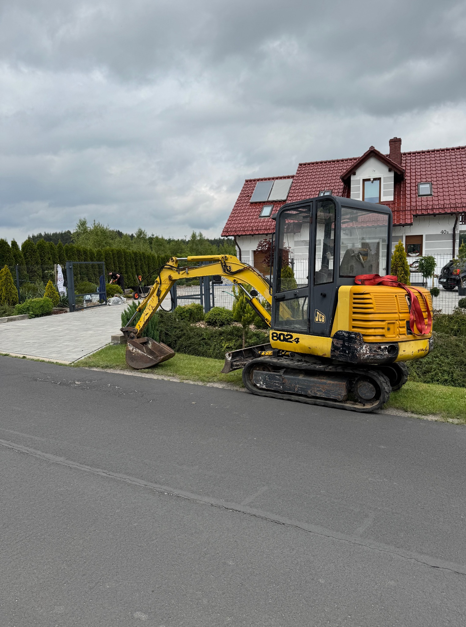 Żółta mini koparka JCB 802.4 stoi na poboczu drogi asfaltowej, w tle dom z czerwoną dachówką i pracownicy przy ogrodzeniu z roślinami. Pochmurne niebo.