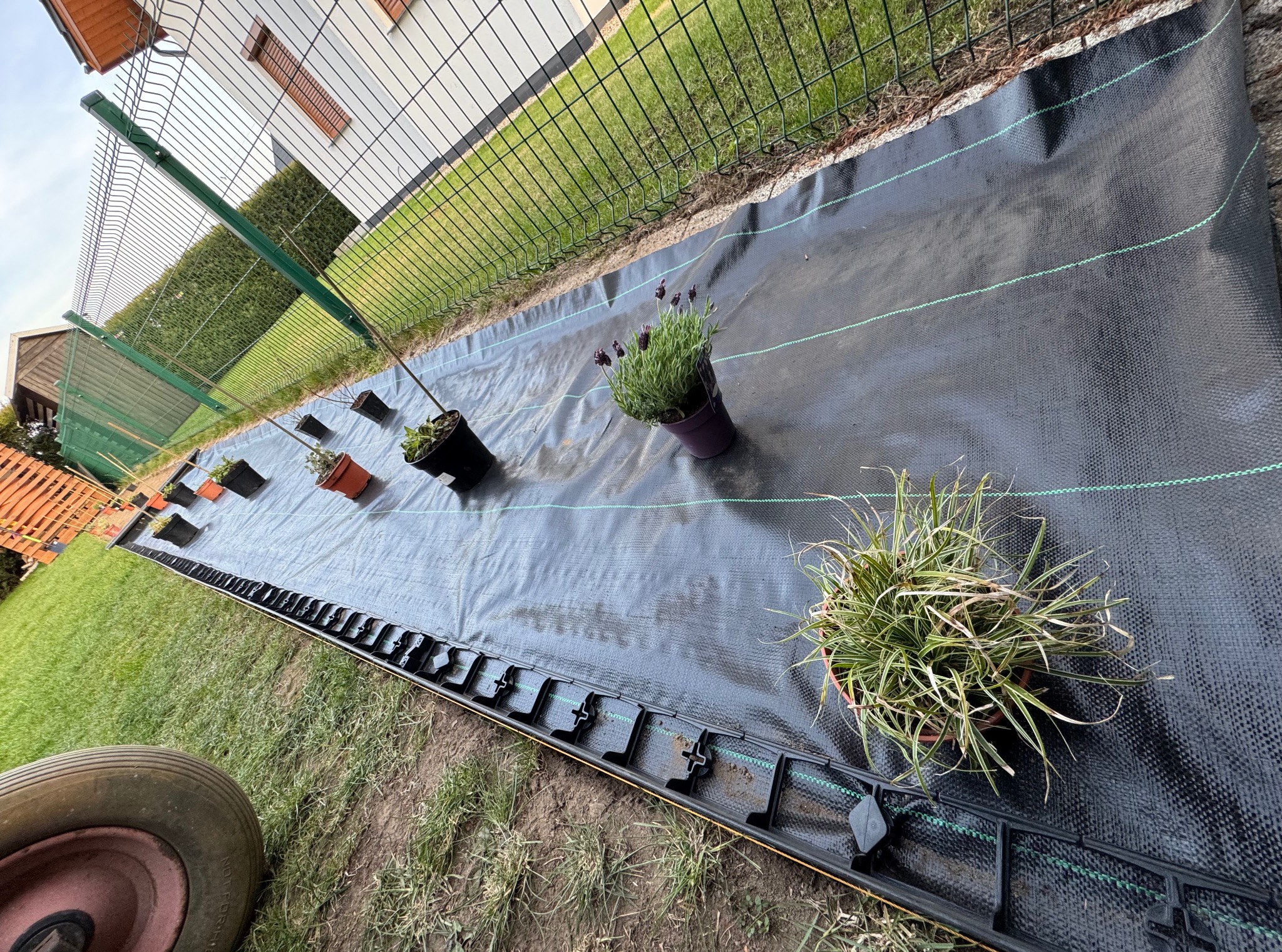 Rozłożona mata szkółkarska z zielonymi liniami, obok doniczki z roślinami i elementy systemu obrzeży trawnikowych. Przygotowanie terenu pod nasadzenia w ogrodzie.