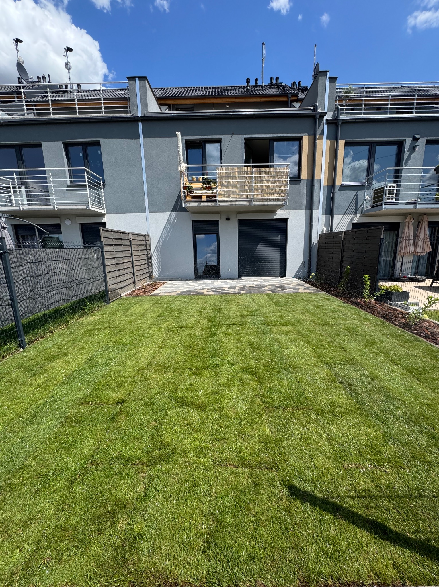 Nowo założony trawnik z rolki przed szeregowcem, z widocznymi elementami systemu nawadniania. Estetyczne wykończenie rabat i ogrodzenia. Słoneczny dzień.