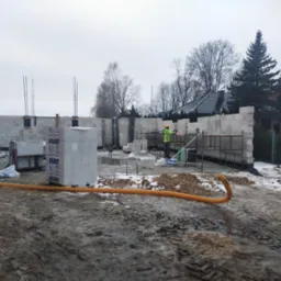 Budowa domu w trakcie: ściany z betonu kom&oacute;rkowego, zbrojenia, pracownik w odblaskowej kamizelce. Na pierwszym planie błotniste podłoże z resztkami śniegu.