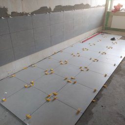 Patryk Cuch - Szare płytki podłogowe w trakcie układania, widoczne krzyżyki dystansowe. Na ścianie ułożone płytki do połowy wysokości. W tle okno z radiatorem.