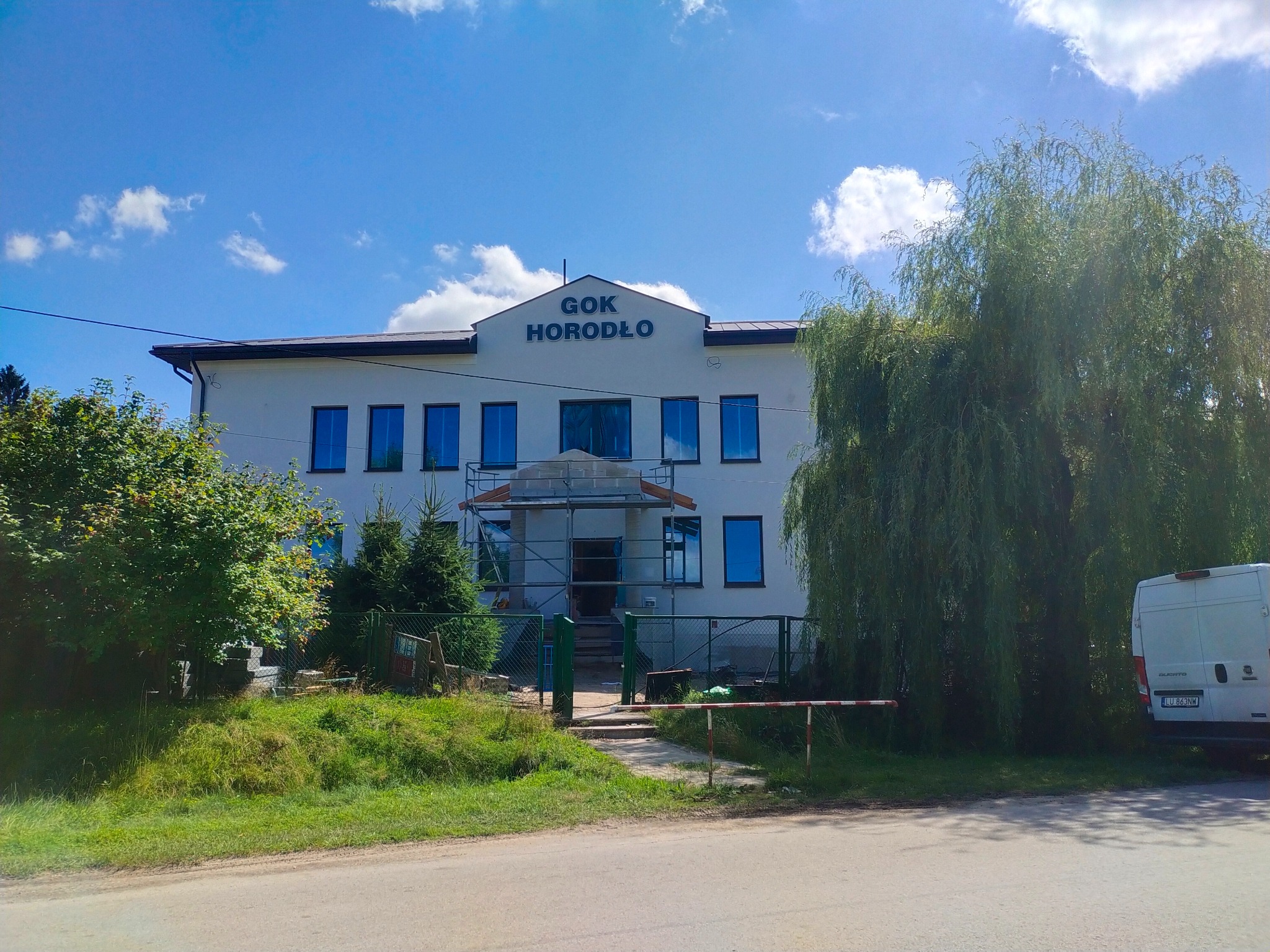 Biały budynek GOK Horodło w trakcie remontu elewacji; widoczne rusztowania przed wejściem, zieleń i fragment białego samochodu dostawczego.