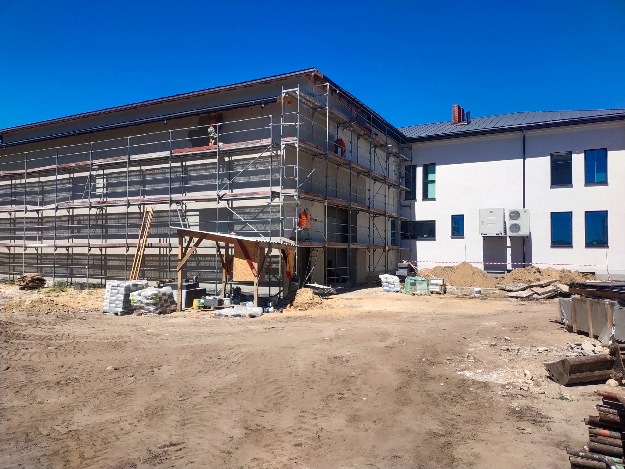 Budynek w trakcie prac elewacyjnych; rusztowania, pracownicy w kamizelkach, worki z zaprawą, teren budowy. Widok na dwukondygnacyjny budynek w słoneczny dzień.