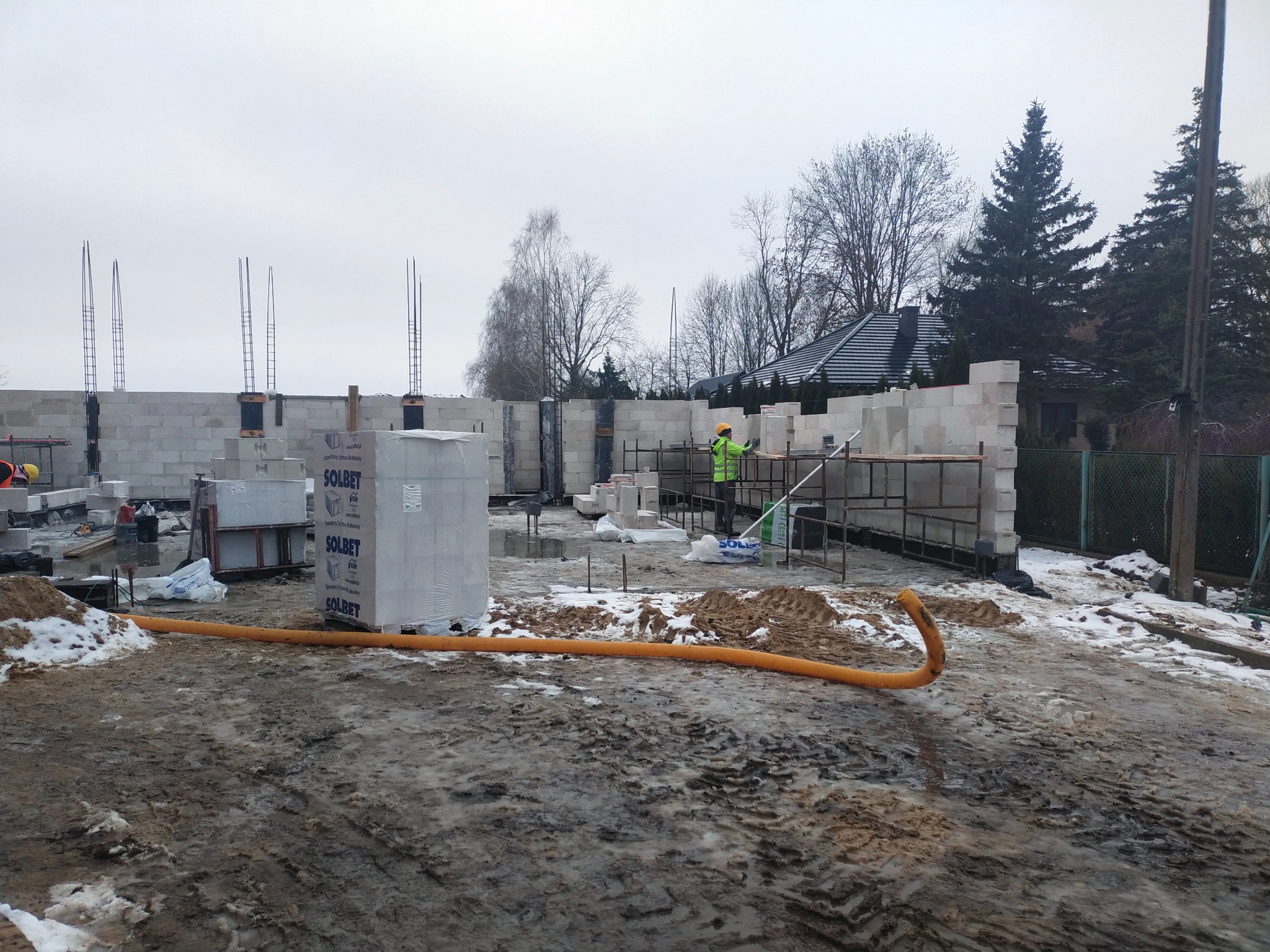 Budowa domu w trakcie: ściany z betonu komórkowego, zbrojenia, pracownik w odblaskowej kamizelce. Na pierwszym planie błotniste podłoże z resztkami śniegu.
