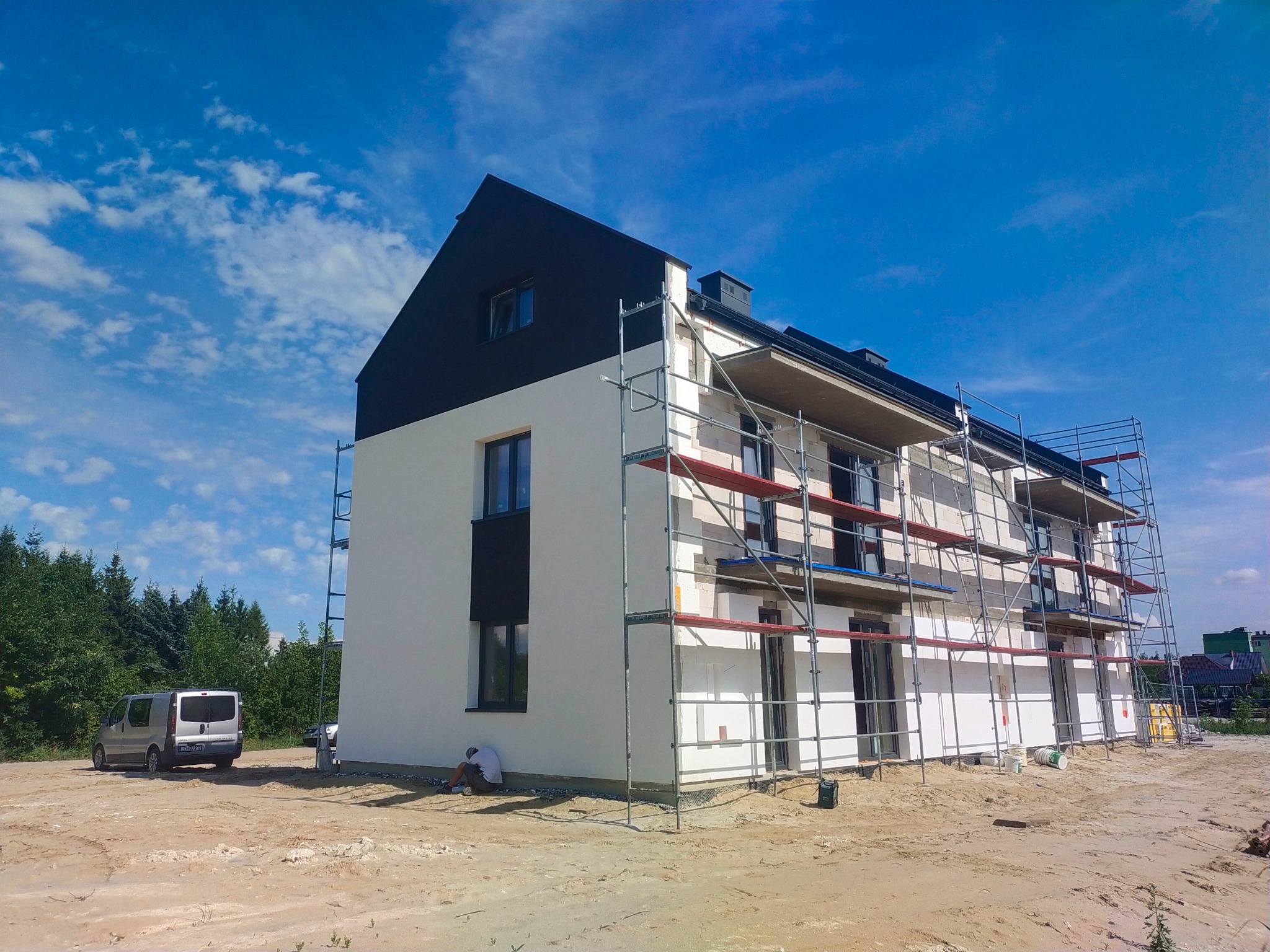 Elewacja dwukondygnacyjnego domu w trakcie prac wykończeniowych; widoczne rusztowania, pracownik przy ścianie, biało-czarna kolorystyka budynku i błękitne niebo.