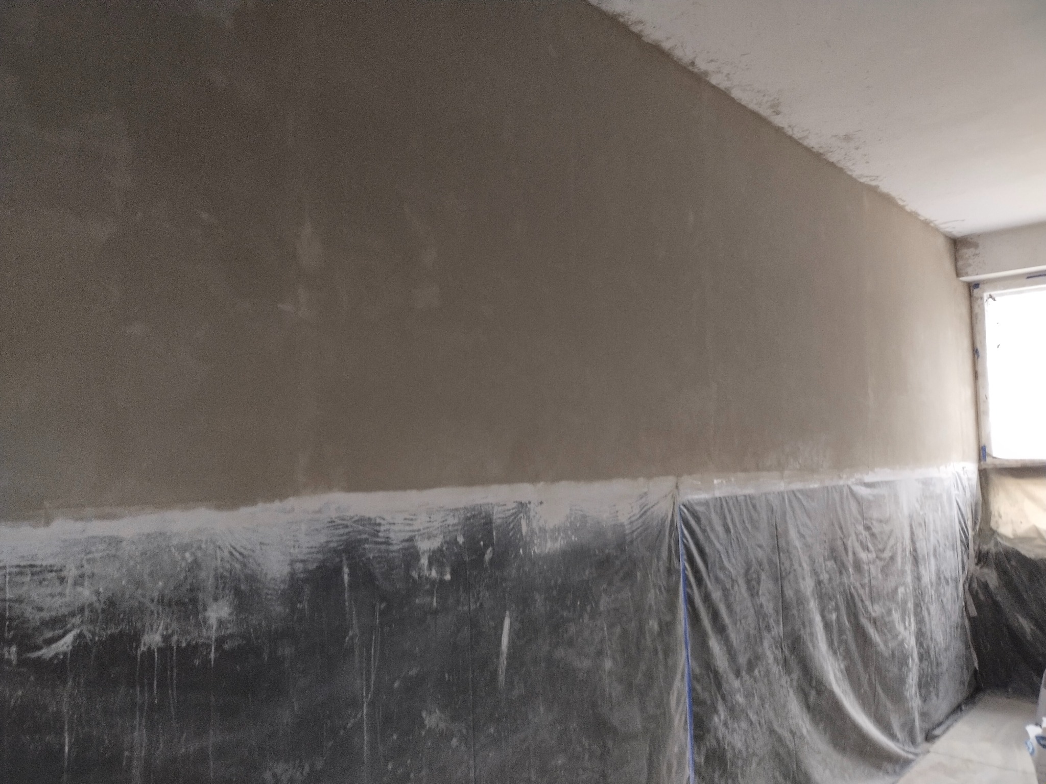 Ściana po szpachlowaniu w Chełmie, zabezpieczona folią malarską. Widoczne nierówności i przygotowanie do dalszych prac wykończeniowych.