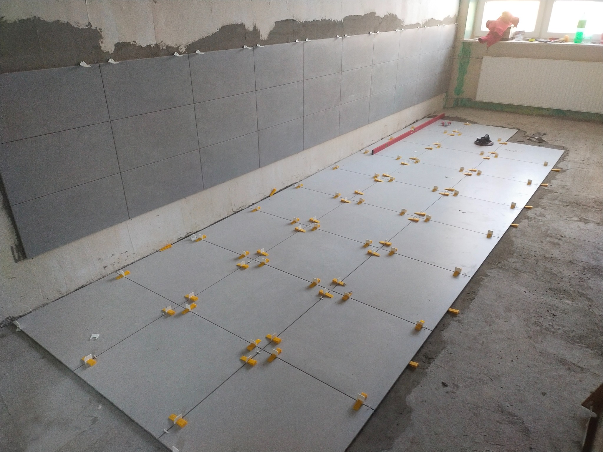 Szare płytki podłogowe w trakcie układania, widoczne krzyżyki dystansowe. Na ścianie ułożone płytki do połowy wysokości. W tle okno z radiatorem.