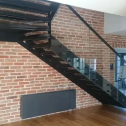 Metalowe schody z szklaną balustradą na tle ceglanej ściany i drewnianej podłogi. Widoczny grzejnik i fragment kolumny. Nowoczesny design.