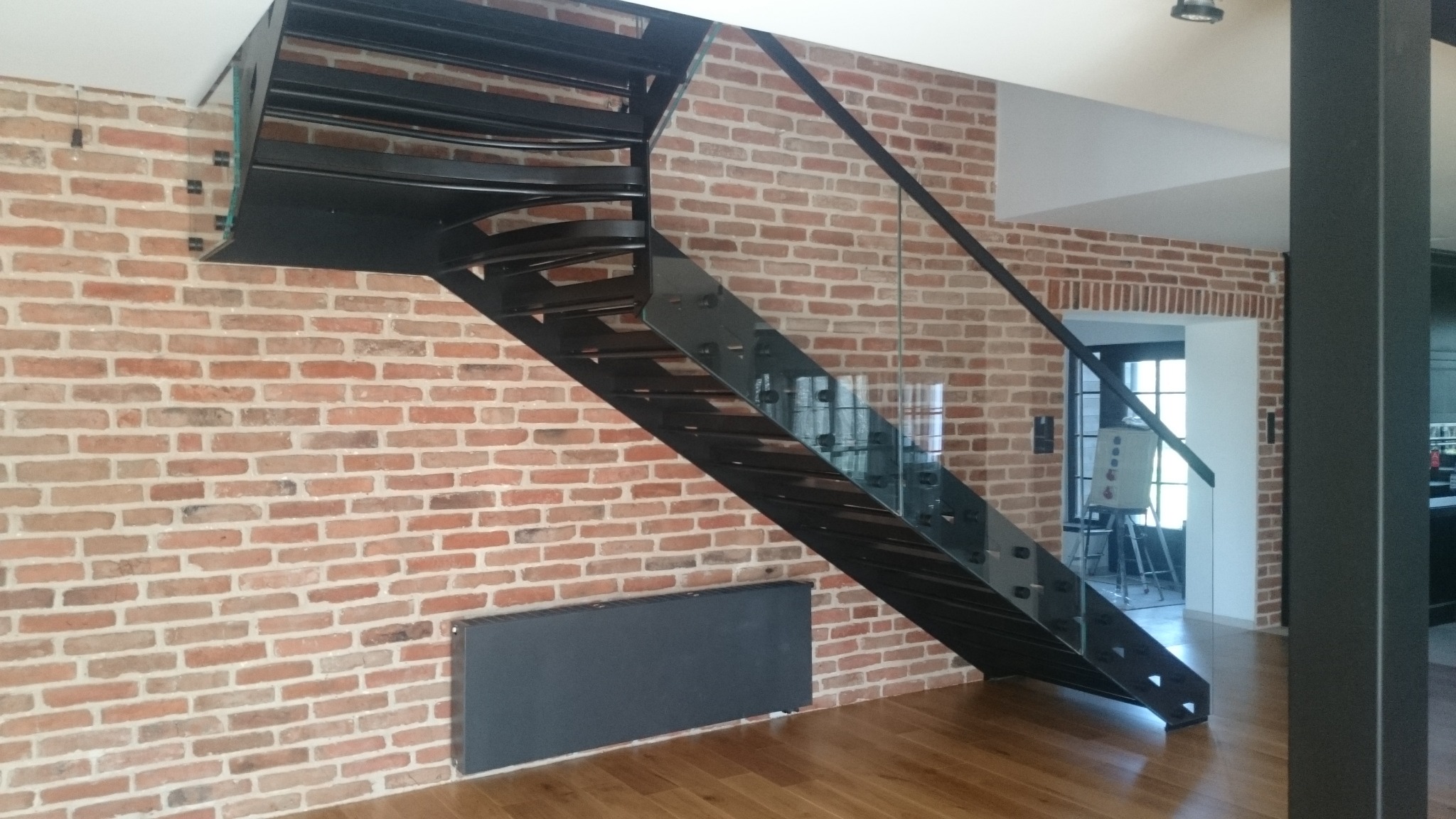 Metalowe schody z szklaną balustradą na tle ceglanej ściany i drewnianej podłogi. Widoczny grzejnik i fragment kolumny. Nowoczesny design.