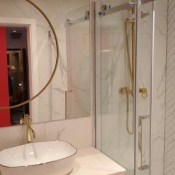 Wykończenia wnętrz - Nowoczesna łazienka z kabiną prysznicową typu walk-in, umywalką nablatową i okrągłym lustrem z złotymi akcentami, marmurowe płytki na ścianach.