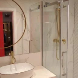 Nowoczesna łazienka z kabiną prysznicową typu walk-in, umywalką nablatową i okrągłym lustrem z złotymi akcentami, marmurowe płytki na ścianach.