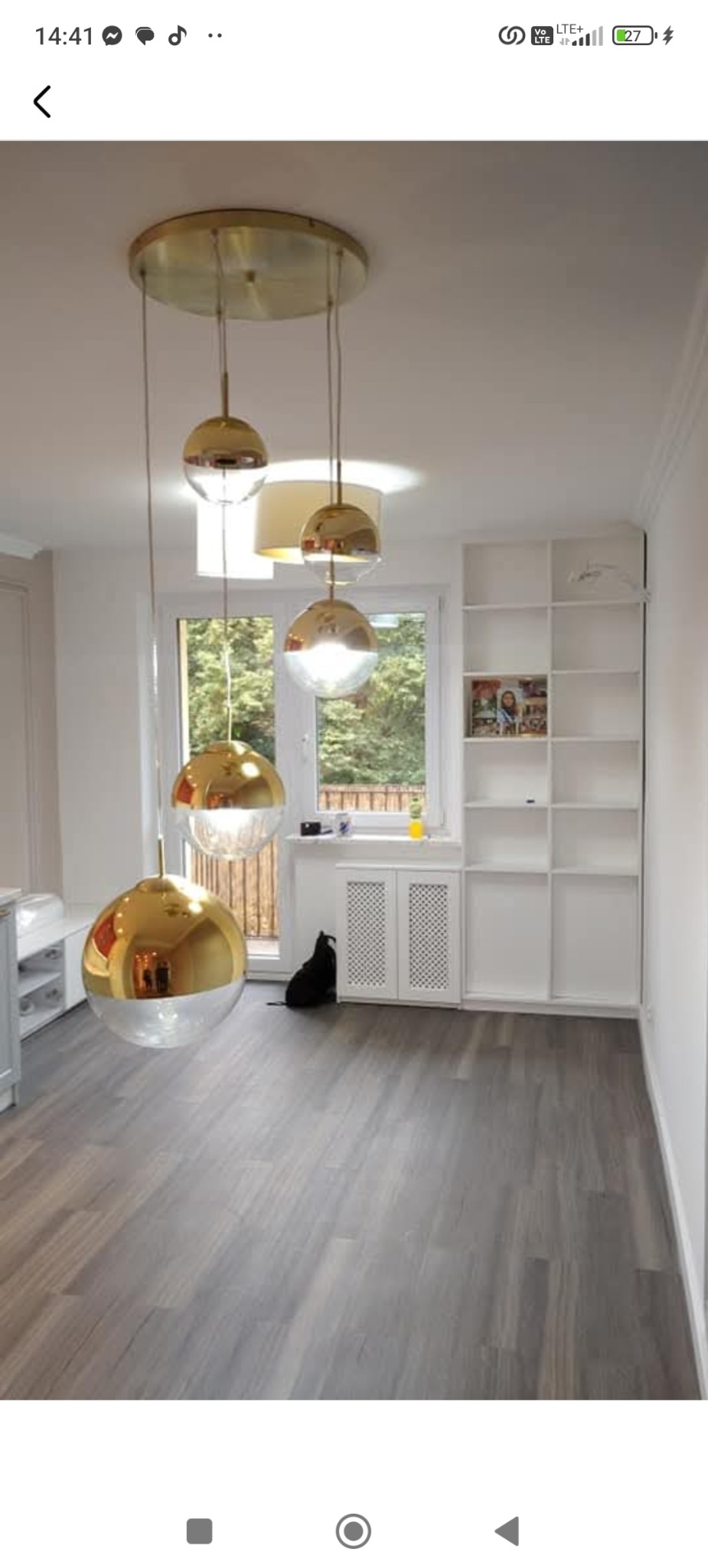 Jasne wnętrze po remoncie z szarą podłogą, białymi ścianami i złotymi lampami wiszącymi. Widoczny fragment okna z zielenią na zewnątrz.