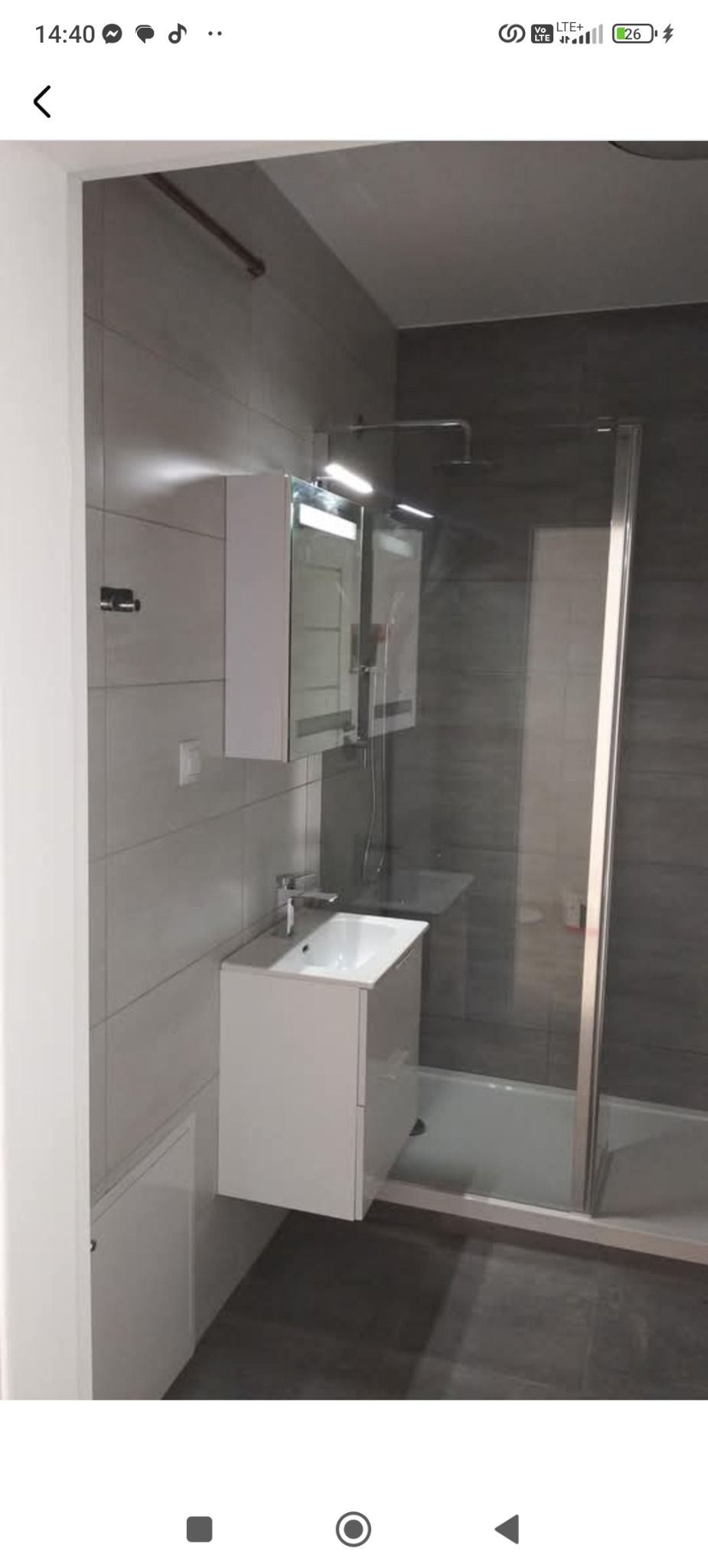 Nowoczesna łazienka po remoncie: szare płytki, biała umywalka z szafką, lustro z oświetleniem, kabina prysznicowa z szklanymi drzwiami i ciemna podłoga.